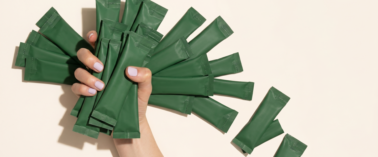 A top-down, clean product shot shows a hand with light-lavender painted fingernails emerging from the bottom center against a plain, light beige background. The hand holds a dense fan of unbranded, green rectangular stick packs or sachets with crimped edges to represent MOQ vs margin skincare pricing and profitability.