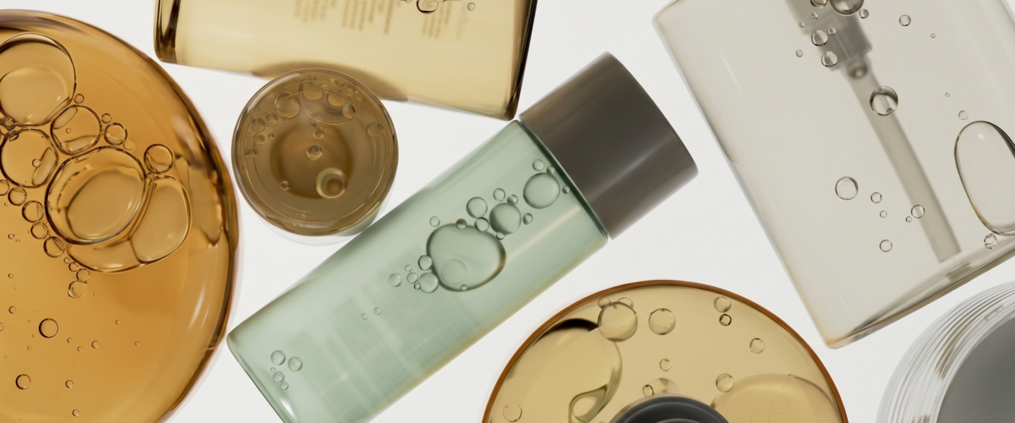 skincare formulation base systems - An overhead macro photograph shows a stylized collection of glass and acrylic cosmetic bottles filled with liquid. At the left is a large, circular vessel with golden-orange oil and prominent, reflective bubbles, partially overlapping with a smaller circular container of the same fluid. Adjacent is a central, cylindrical bottle filled with pale green oil and a cluster of suspended bubbles. To the right, a clear bottle reveals a dip tube and individual air bubbles, while a grey-capped bottle is positioned below it and above another container of golden-orange liquid. The white background emphasizes the translucent textures, varying colors, and geometric shapes of the containers, creating a sophisticated and clean visual composition.