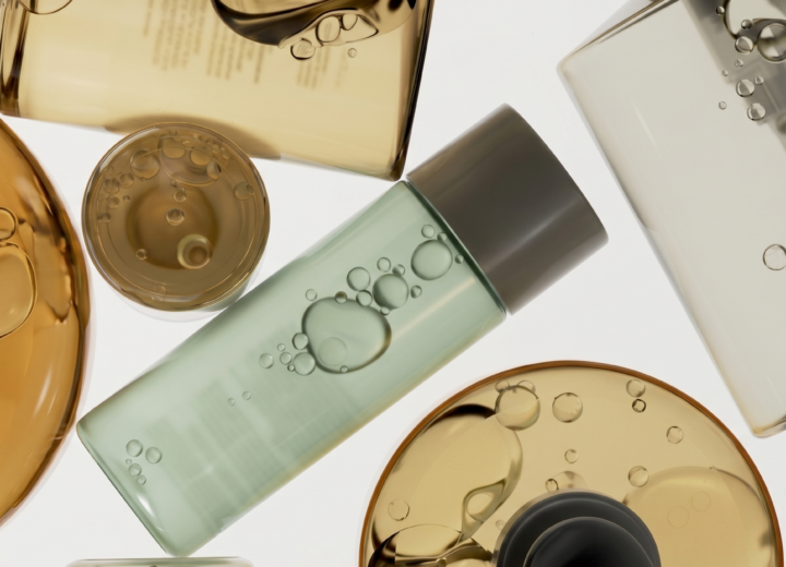 skincare formulation base systems - An overhead macro photograph shows a stylized collection of glass and acrylic cosmetic bottles filled with liquid. At the left is a large, circular vessel with golden-orange oil and prominent, reflective bubbles, partially overlapping with a smaller circular container of the same fluid. Adjacent is a central, cylindrical bottle filled with pale green oil and a cluster of suspended bubbles. To the right, a clear bottle reveals a dip tube and individual air bubbles, while a grey-capped bottle is positioned below it and above another container of golden-orange liquid. The white background emphasizes the translucent textures, varying colors, and geometric shapes of the containers, creating a sophisticated and clean visual composition.