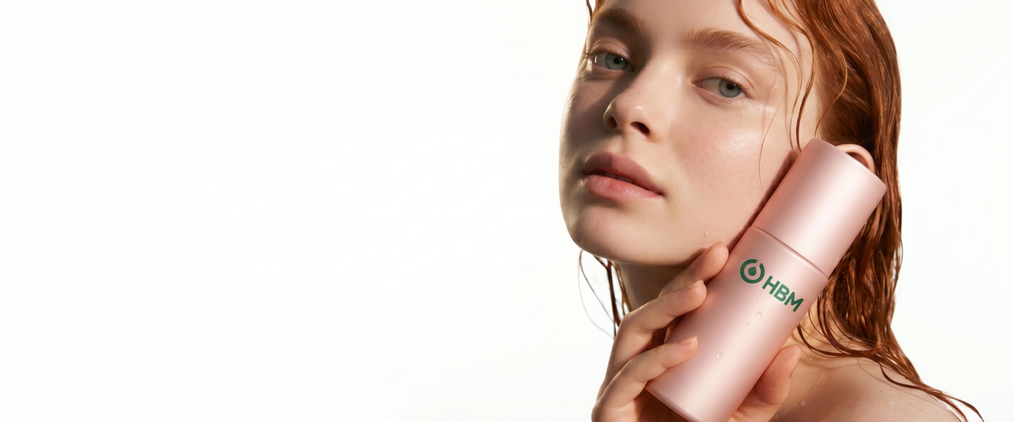 Gel vs Cream vs Serum Bases: A close-up studio photograph of a young woman with wet, reddish-auburn hair and radiant skin, looking directly at the camera. She is holding a metallic pink cosmetic bottle of serum with the green "HBM" logo against her cheek, set against a plain white background.