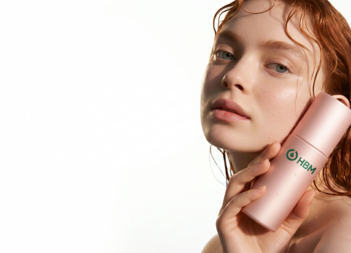 Gel vs Cream vs Serum Bases: A close-up studio photograph of a young woman with wet, reddish-auburn hair and radiant skin, looking directly at the camera. She is holding a metallic pink cosmetic bottle of serum with the green "HBM" logo against her cheek, set against a plain white background.